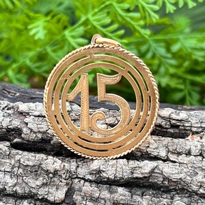 May include: Gold-toned pendant with the number 15 in the center, surrounded by a circular frame with a rope-like border. The pendant is displayed against a textured wooden surface with green foliage in the background.