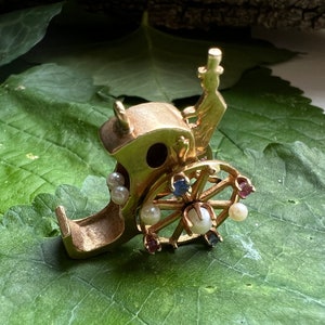 May include: A gold charm featuring a miniature Ferris wheel with colorful gemstones and white pearls. The charm is designed in the shape of a carriage with a small figure sitting on top.