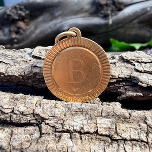 May include: A circular gold-toned metal pendant featuring a central engraved letter B. The pendant has a textured, radiating sunburst pattern around the outer edge and includes a small circular jump ring at the top for attachment to a chain.