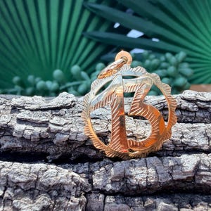May include: A circular gold pendant featuring the number 13 in the center. The pendant has a scalloped edge and a textured, engraved finish on the surface of the number and the outer border, with a small loop at the top for attachment.