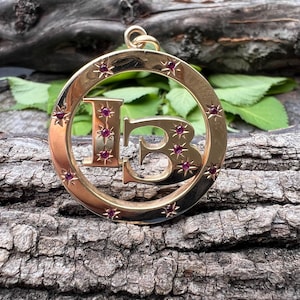 May include: Gold-toned pendant featuring the letters "I" and "B" within a circular frame. The pendant is adorned with small, star-shaped accents, each containing a red gemstone. The pendant is suspended from a gold loop.