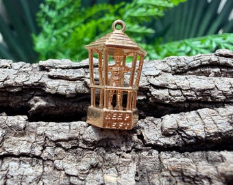 May include: A gold-toned metal charm shaped like a hexagonal lantern or kiosk. Inside the structure stands a small figure of a person. The base of the charm features the embossed text KEEP LEFT. It includes a small loop at the top for attachment.