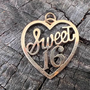 May include: A gold-colored pendant in the shape of a heart. The words "Sweet 16" are written in a cursive font within the heart. The pendant has a loop at the top for a chain.