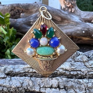 May include: A gold pendant charm in the shape of a square with a basket filled with colorful gemstones. The gemstones include green, blue, red, and white.