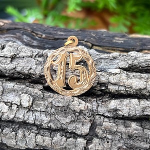 May include: Gold-colored pendant with the number 15 in a decorative font, encircled by a floral design. The pendant has a loop at the top for a chain. The background is a textured, dark brown wooden surface.