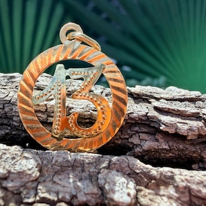 May include: A circular gold pendant featuring the number 13 in the center. The outer ring has a textured, diagonal striped pattern, while the number 13 is detailed with small, raised dots along its edges. A small gold loop is attached at the top.
