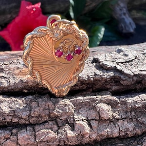 May include: A gold-colored pendant with a heart-shaped design, featuring a sunburst pattern and three small red gemstones. The pendant is set against a natural wood background with a red flower in the background.