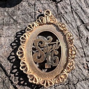 May include: Gold-toned metal pendant with a decorative oval frame and the words "Sweet 16" in a stylized font.