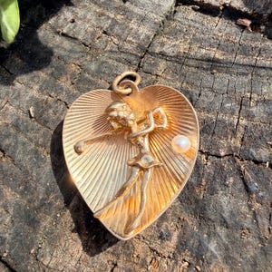 May include: A gold-colored heart-shaped pendant featuring a golfer in mid-swing. The pendant has a textured, sunburst design and a small pearl accent. The golfer is depicted with detailed features, and the pendant has a loop for a chain.