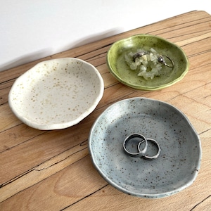 Puede incluir: Tres platos de cerámica para joyas en diferentes colores. Los platos están moteados de marrón y blanco, verde y blanco, y azul y blanco. Los platos están sobre una superficie de madera.