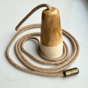 May include: A ceramic light pull featuring a two-tone design with a glossy tan glazed top and a matte cream base. The pull is attached to a long, braided tan cord that ends in a cylindrical, antique-style brass connector piece for easy installation.