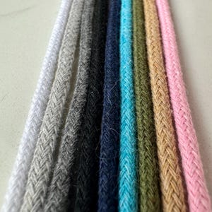 Pode incluir: Uma coleção de oito cordões trançados paralelos dispostos verticalmente, apresentando um espectro de cores incluindo branco, cinza claro, cinza escuro, preto, azul marinho, turquesa brilhante, verde oliva, bege e rosa claro com trama texturizada.