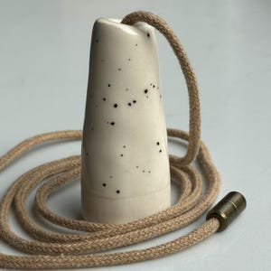 May include: A light cream-coloured ceramic light pull with dark brown speckles, featuring a tapered cylindrical shape. A braided tan cord runs through the top of the ceramic piece and includes a small, cylindrical antique brass-toned metal connector.