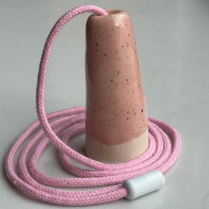 Può includere: Un lampadario in ceramica rosa con un cavo intrecciato rosa abbinato. Il lampadario è a forma di cono con una finitura maculata e una base bianca. Un connettore cilindrico bianco è all'estremità del cavo.