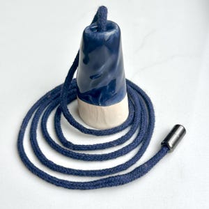 May include: A ceramic light pull with a glossy dark blue glazed top and a matte off-white base. A long navy blue braided cord is threaded through the centre of the pull, coiled underneath, and finished with a small, dark metallic cylindrical connector.
