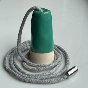 Op de afbeelding: Een cilindrische lichtschakelaar met een glanzend groenblauw geglazuurd bovendeel en een matte, gebroken witte, ongeglazuurde basis. Een grijs gevlochten koord loopt door het keramische onderdeel en eindigt in een gepolijste zilveren metalen connector.