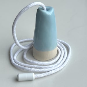 Op de afbeelding: Een lichtblauwe keramische trekschakelaar met een gladde geglazuurde afwerking op de bovenste helft en een matte, ongeglazuurde beige basis. De trekker is bevestigd aan een lang, wit gevlochten koord met een kleine, cilindrische witte plastic connector aan het uiteinde.