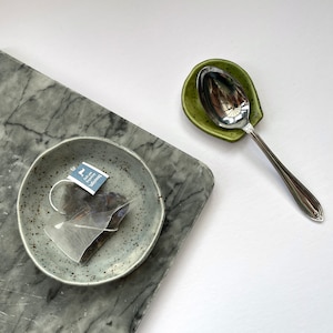 May include: A speckled light gray ceramic dish containing a triangular teapigs earl grey tea bag, a small speckled olive green ceramic spoon rest, and a polished silver metal spoon. The items are arranged on a gray marble surface.