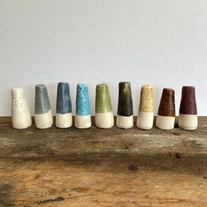Op de afbeelding: Tien keramische vazen in verschillende kleuren, waaronder blauw, groen, bruin en wit. De vazen zijn allemaal even groot en hebben dezelfde vorm, en ze zijn in een rij op een houten oppervlak gerangschikt.