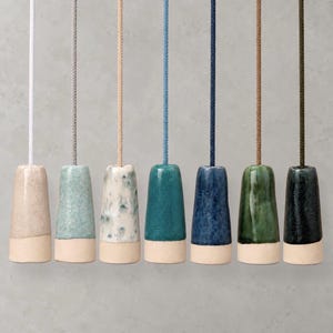 May include: Seven ceramic light pulls with attached cords, each featuring a tapered cylindrical shape. The pulls display various glazed finishes in shades of beige, light blue, speckled white, teal, dark blue, green, and black, with matte bases.