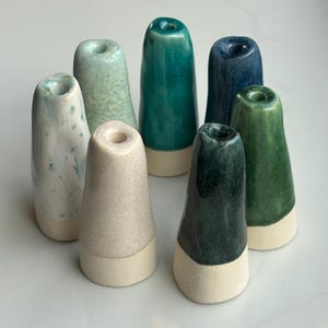 Op de afbeelding: Een verzameling van zeven keramische flesstoppers in verschillende kleuren, waaronder teal, groen en blauw. Elke stopper heeft een conische vorm met een witte basis en een gat aan de bovenkant. De stoppers zijn dicht bij elkaar geplaatst en tonen hun diverse tinten en texturen.