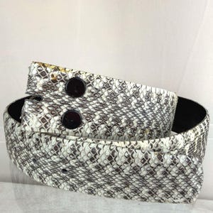 May include: A white and grey snakeskin belt with two large, dark brown buttons. The belt is folded over itself, showing the black underside. The snakeskin pattern is detailed and textured.