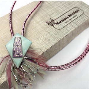 May include: A bolo tie with a light blue diamond-shaped pendant featuring a purple floral design. The pendant is adorned with feathers and beads. The cord is a shiny pink. The box in the background has the text "Mariposa Boutique, Taos, New Mexico."