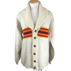May include: A cream-colored knit cardigan featuring a wide shawl collar and a front button closure with five brown textured buttons. The garment displays a horizontal striped pattern across the chest in shades of orange, red, and dark maroon.