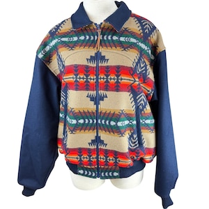 May include: A zip-up jacket featuring a tan center panel with a multi-colored geometric pattern in red, orange, green, and navy blue. The sleeves, collar, and ribbed hem are solid navy blue. The jacket has a full-length front zipper with a leather pull.
