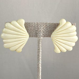 May include: A pair of off-white seashell-shaped earrings. Each earring features a fan-like design with multiple curved ridges, resembling a scallop shell. The earrings are displayed on a neutral-colored earring stand.