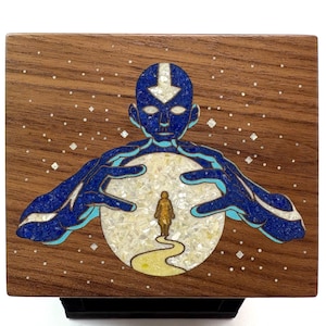 Op de afbeelding: Houten doos met een mozaïekontwerp. Het toont een blauwe figuur met uitgestrekte handen rond een centrale cirkel met een kleinere figuur. De achtergrond is een sterrenhemel, met een bruin houten frame.