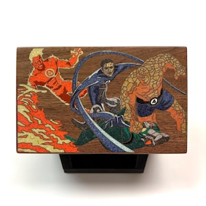 May include: A rectangular wooden box lid featuring an inlaid graphic of the Fantastic Four characters. The design displays a figure engulfed in orange flames, a blue-suited character with elongated limbs, and a rocky orange figure in blue shorts.