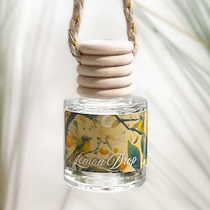 May include: A small, clear glass bottle with a light-colored wooden cap and a hanging rope. The label features a yellow and green botanical illustration with a bird and the text Lemon Drop printed in a white script font on the front.