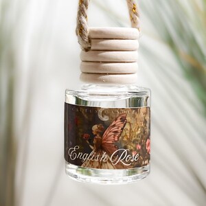 May include: A small, clear glass bottle hanging from a thick, twisted beige rope. The bottle features a wooden cap and a rectangular label with a dark, detailed illustration of a fairy with wings and the text English Rose printed in white.