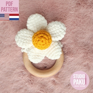 Op de afbeelding: Een witte gehaakte bloem met een geel centrum bevestigd aan een houten bijtring. De ring ligt op een roze pluche oppervlak. De tekst "STUDIO PAKU CROCHET DESIGNS" is zichtbaar in de rechterbenedenhoek van de afbeelding.