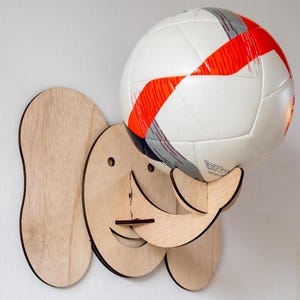 Op de afbeelding: Een houten, olifantvormige voetbalhouder. De houder is gemaakt van lichtgekleurd hout en heeft een lachend gezicht en grote oren. Een witte voetbal met rode en grijze strepen rust in de houder.