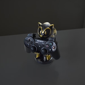 Puede incluir: Una figura negra y dorada de una criatura sosteniendo un mando negro de Sony PlayStation. El mando tiene la palabra "SONY" impresa. La figura está sobre una superficie reflectante.