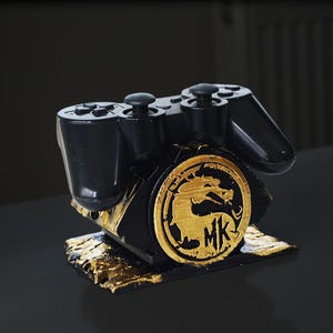 Pode incluir: Um suporte para controlador de videogame impresso em 3D preto e dourado com um logotipo de dragão Mortal Kombat.