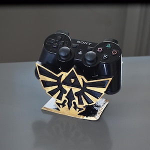 Può includere: Un controller PlayStation nero è posizionato su un supporto dorato con un design Triforce nero e oro. Il supporto è in plastica e ha una superficie liscia.