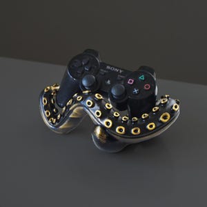 Puede incluir: Un soporte en forma de pulpo negro y dorado sostiene un mando de PlayStation negro. El mando tiene la palabra "SONY" impresa en él.