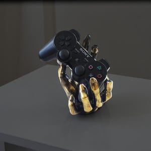 May include: A black PlayStation controller is held by a gold-colored, sculpted hand. The controller has the word "SONY" printed on it.