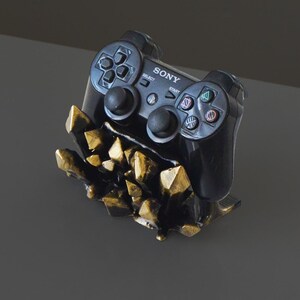 Puede incluir: Un mando de videojuegos negro se encuentra en un soporte de cristal de color dorado. El mando tiene la palabra "SONY" impresa en él.