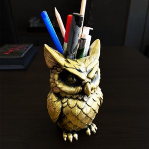 Könnte beinhalten: Ein goldfarbener, eulenformiger Schreibtisch-Organizer mit Stiften, Bleistiften und Markern. Die Eule hat detaillierte Federstrukturen und einen strengen Ausdruck. Der Organizer besteht aus einem metallischen Material und steht auf einer dunklen Oberfläche.