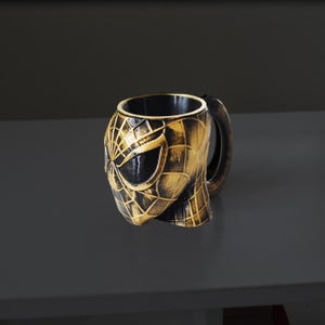 Puede incluir: Una taza negra y dorada con temática de Spider-Man. La taza tiene la forma de la máscara de Spider-Man, con un diseño de telaraña texturizada y un interior negro. El asa es de color marrón oscuro.