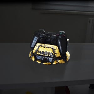 May include: A black Sony PlayStation controller rests on a custom stand. The stand is gold and black with the name "MARIO" in white lettering. The controller is black with the Sony logo.