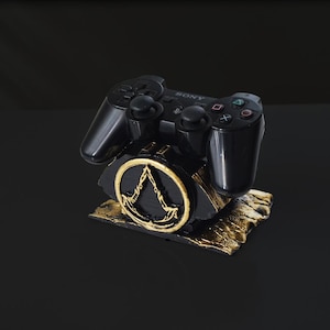 May include: A black PlayStation controller sits on a black and gold stand with the Assassin's Creed logo. The stand is made of a textured material and has a gold-colored finish.