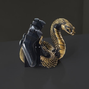 Op de afbeelding: Een decoratieve boekensteun in de vorm van een cobra in goud en zwart. De slang is opgerold, met zijn kop omhoog, en houdt een zwarte videogamecontroller vast. De boekensteun is ontworpen om boeken of andere items vast te houden.