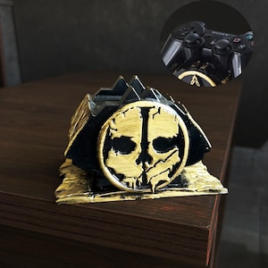 May include: A black and gold Call of Duty-themed controller stand. The stand features a skull design on a circular emblem. The stand is on a dark brown surface. A close-up of a black controller is in the upper right corner.