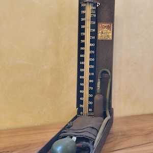 Puede incluir: Un antiguo monitor de presión arterial con una caja de metal oscuro. El medidor tiene una escala en blanco y negro con marcas azules y amarillas. Incluye una pera, tubos y un manguito de tela. La etiqueta dice "HYEON".