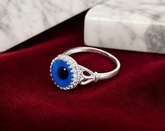 May include: A silver ring featuring a circular evil eye charm with a bright blue iris and a black pupil. The setting has a scalloped edge with small metallic beads, and the band includes an openwork design on the sides near the central charm.